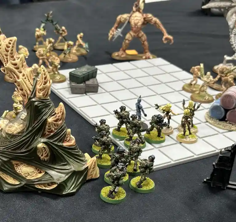 A photo of a tabletop wargame setup shows miniature soldiers in green tactical gear facing off against monstrous alien…