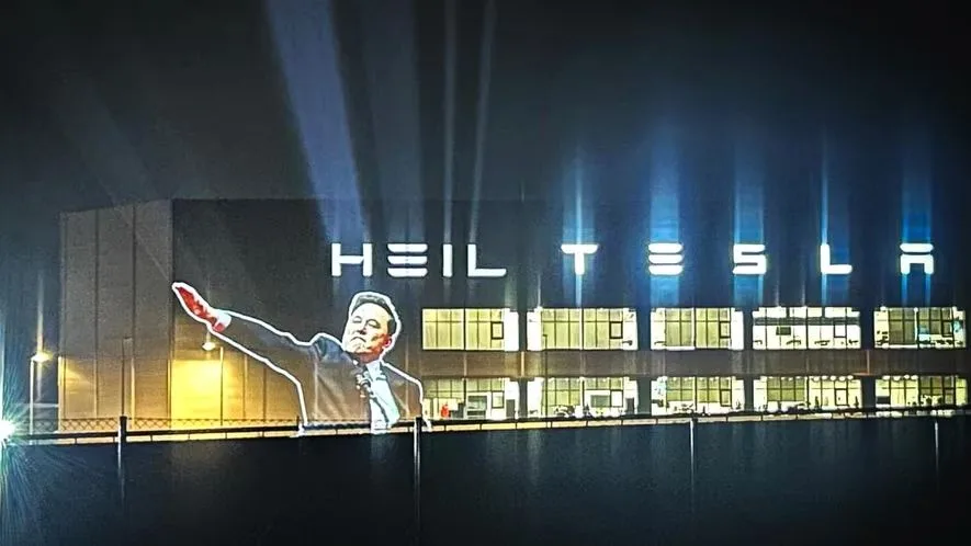 A nighttime photo of a large building with illuminated windows and spotlights. Projected onto the building's facade is a…