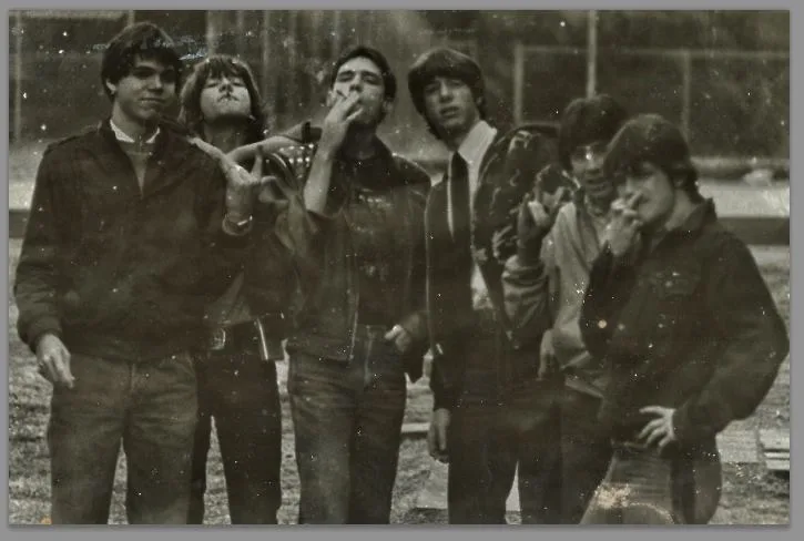 A grainy, black-and-white photograph of six young men, likely the Rolling Stones, standing outdoors in vintage attire…