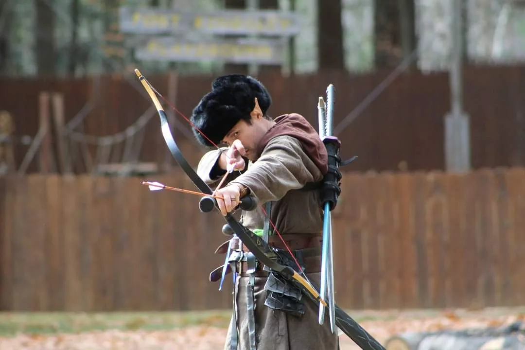 A man dressed in historical reenactment attire, including a fur hat and brown tunic, is aiming a longbow with intense focus…