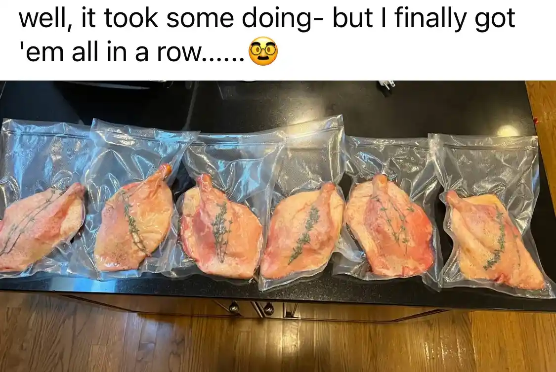 Six vacuum-sealed packages of raw poultry, each with a sprig of thyme, are lined up neatly on a black countertop. The…
