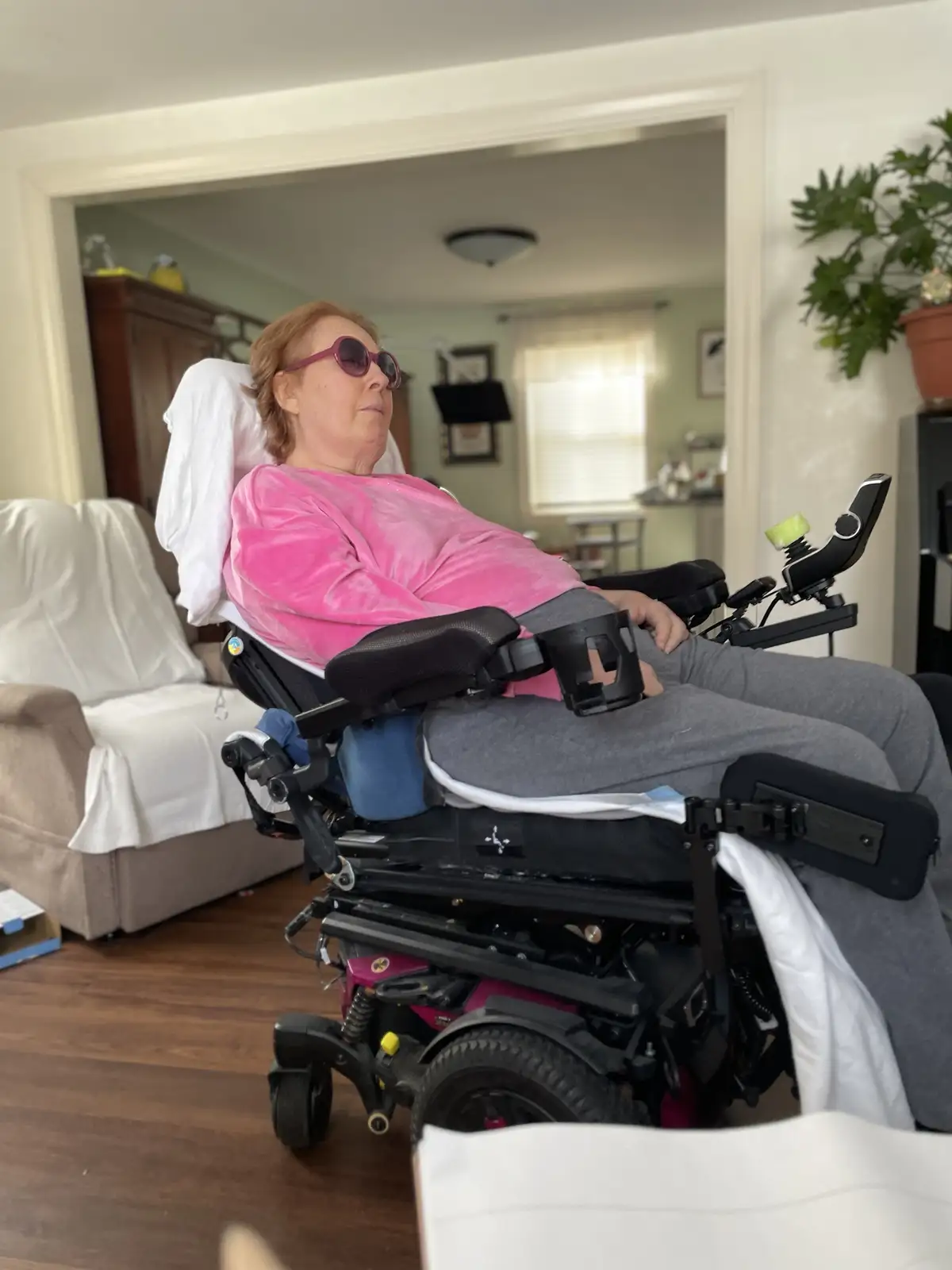 A woman with reddish hair wearing pink sunglasses and a pink shirt sits in a motorized wheelchair indoors, looking relaxed…