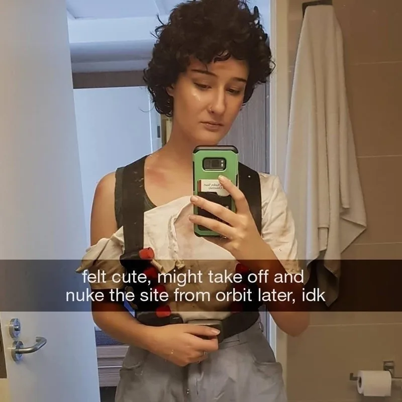 A person with short curly hair takes a mirror selfie in a bathroom while wearing a white shirt and grey pants, holding a…