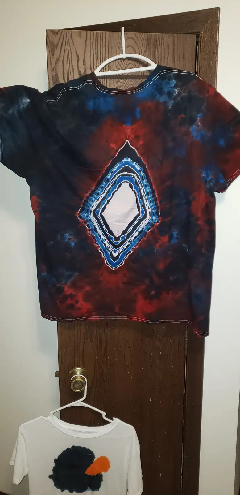 A tie-dye t-shirt with a blue and red abstract design hangs on a white hanger on a wooden door, with a smaller white t-shirt…