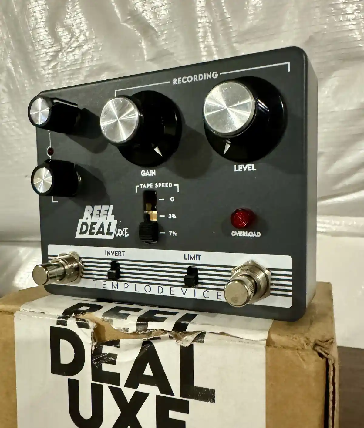 A black guitar effects pedal labeled 'REEL DEAL LUXE' by 'TEMPLODEVISE' sits atop a cardboard box with the same name. The…