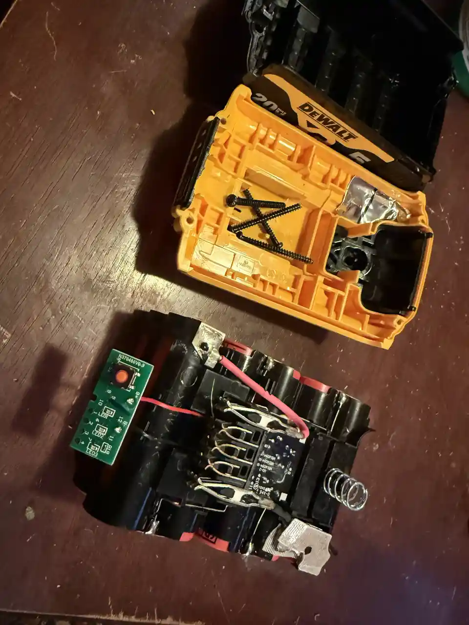 An image shows a disassembled DeWalt power tool battery lying on a wooden surface, with its orange plastic casing open and…