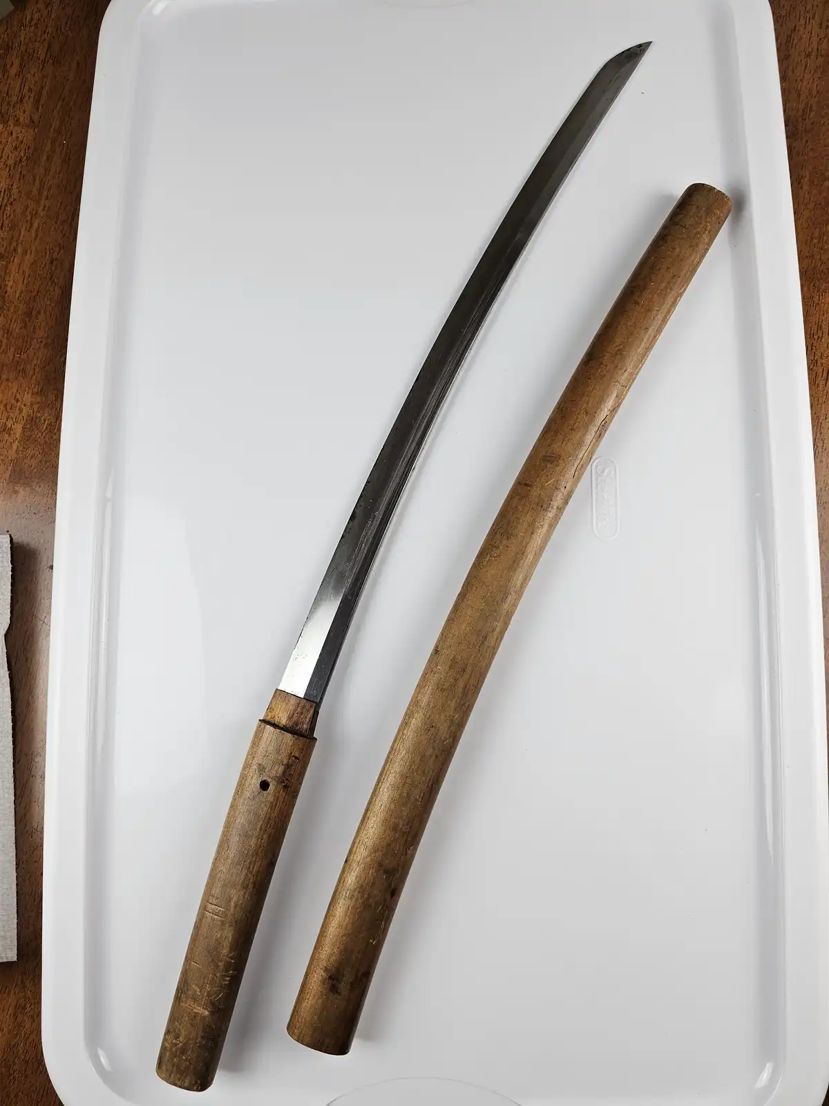 A curved, metallic knife with a wooden handle lies next to its matching wooden sheath on a white plastic tray, which has a…