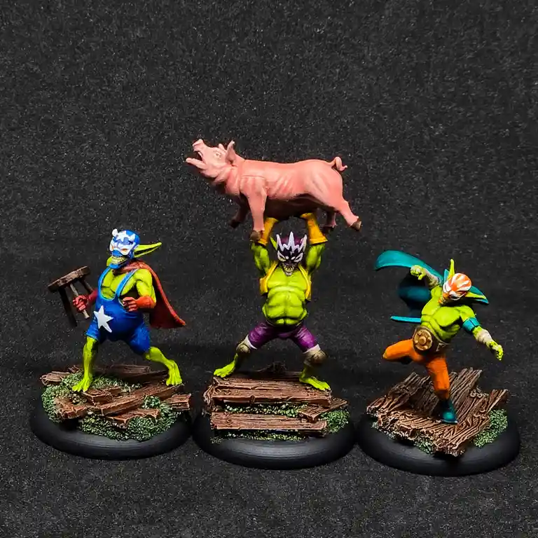 Three painted miniature figures resembling luchador-style superheroes stand on wooden bases against a dark background. The…
