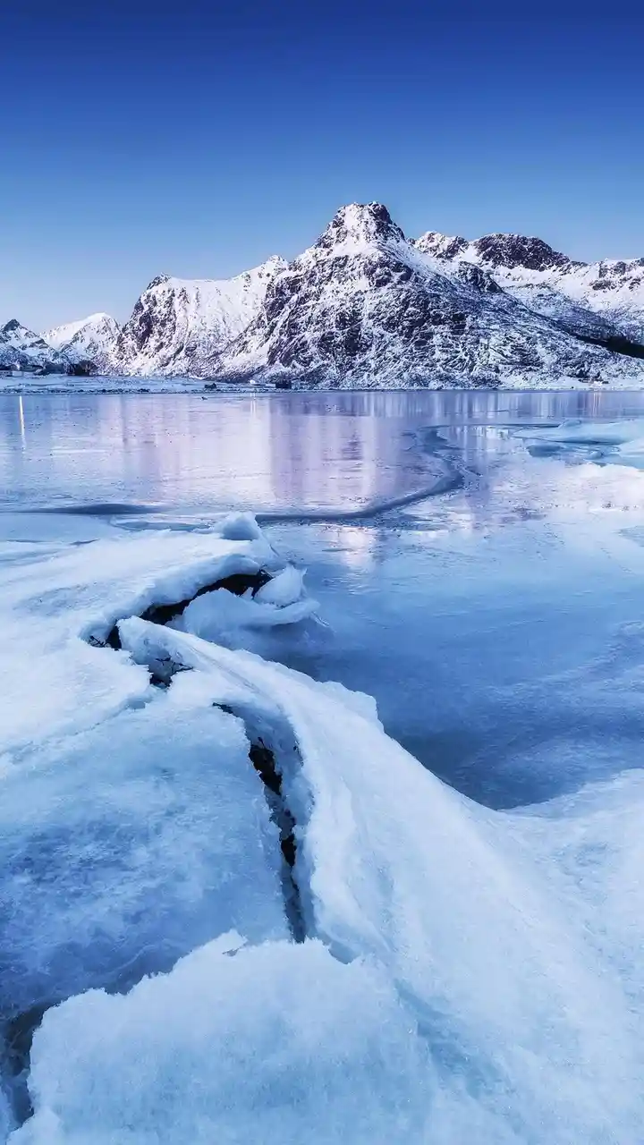 A serene, icy landscape featuring snow-covered mountains reflected in a frozen lake under a clear blue sky, with a prominent…