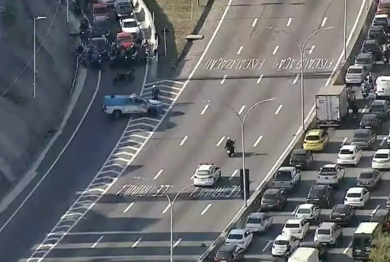 An aerial view of a multi-lane highway with heavy traffic and police vehicles. A lone motorcyclist rides down the center…