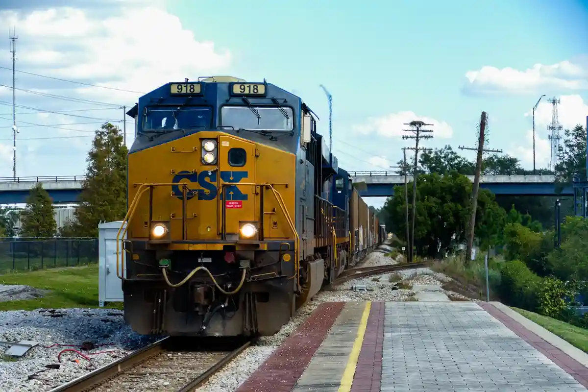 A CSX freight train, numbered 918, is shown approaching a station platform under a partly cloudy sky. The train's front has…