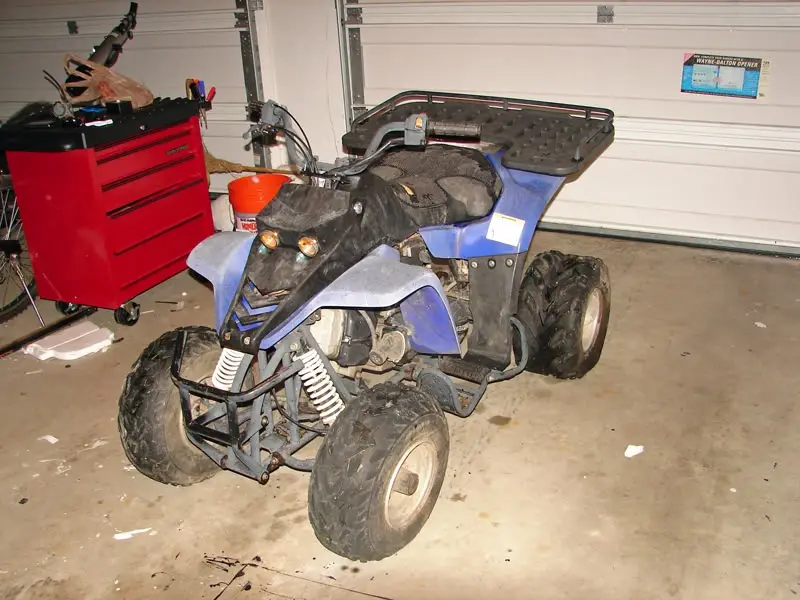 A blue and black four-wheeler ATV sits in a cluttered garage next to a red tool chest. The vehicle appears heavily modified…