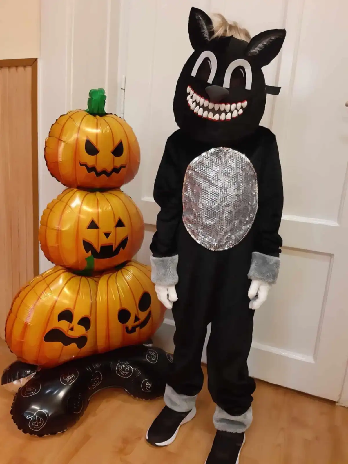 A child wearing a black Halloween costume with a large, cartoonish cat mask featuring white eyes and sharp teeth stands next…