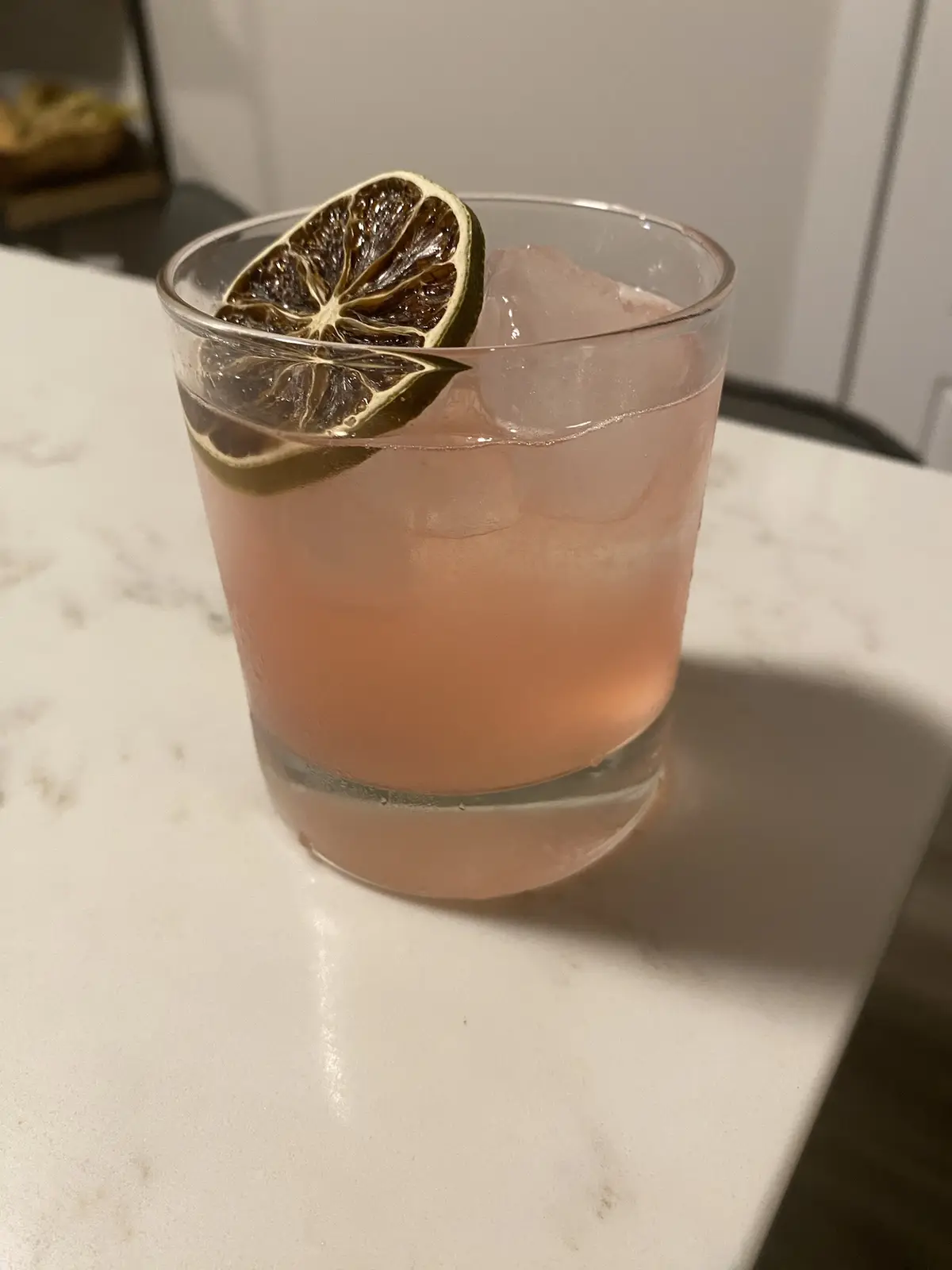 A close-up photo shows a pink cocktail in a rocks glass with ice and a dried lime wheel garnish on the rim, sitting on a…