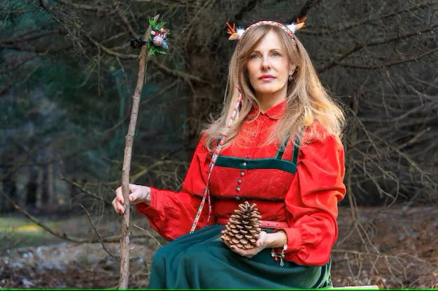 A woman with long blonde hair, wearing a red blouse and green skirt, sits in a forest holding a pinecone in one hand and a…