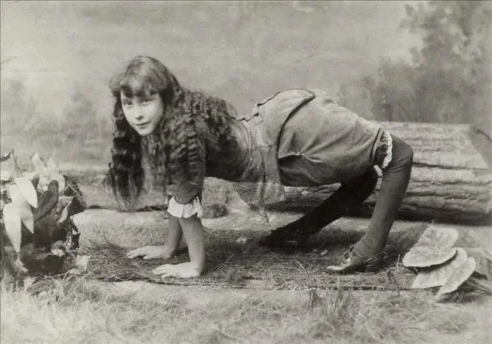 A vintage black-and-white photograph shows a young woman with long hair in a posed, low, crawling position on the ground…