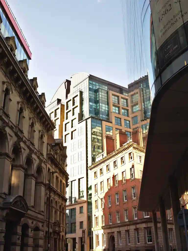 A low-angle urban photo shows a mix of old ornate stone buildings and modern glass skyscrapers under a clear blue sky. A…