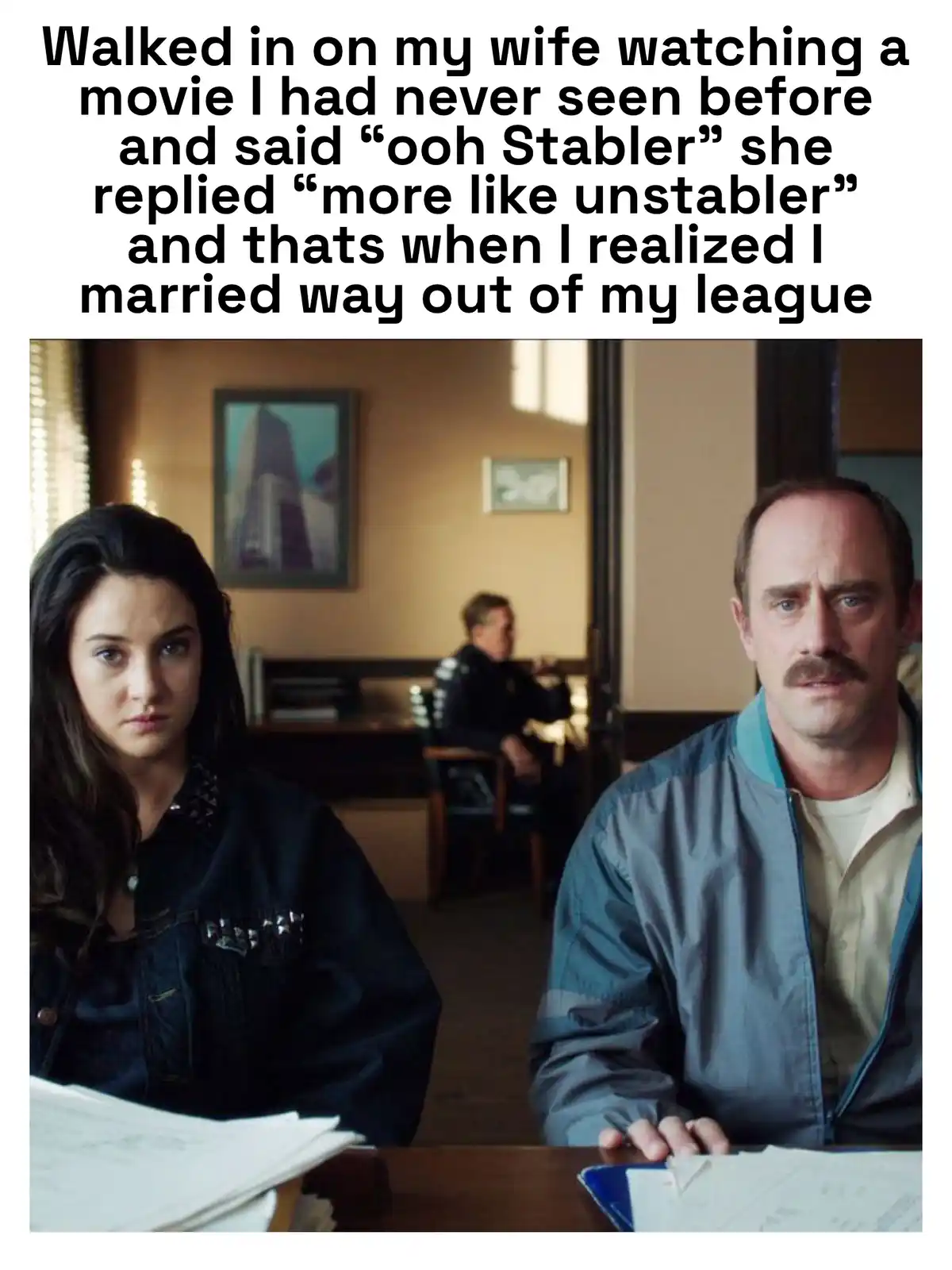The image shows a man and a woman sitting at a table, both looking directly at the camera with serious expressions. The man…