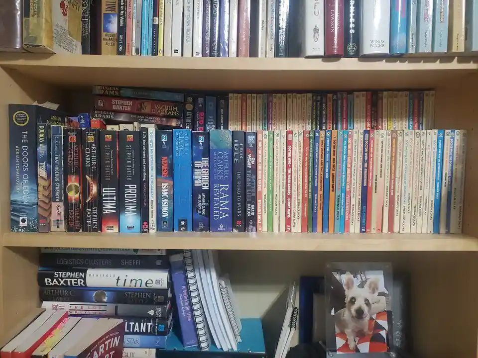 A bookshelf filled with science fiction novels, including many Doctor Who books, with a small photo of a dog placed on the…