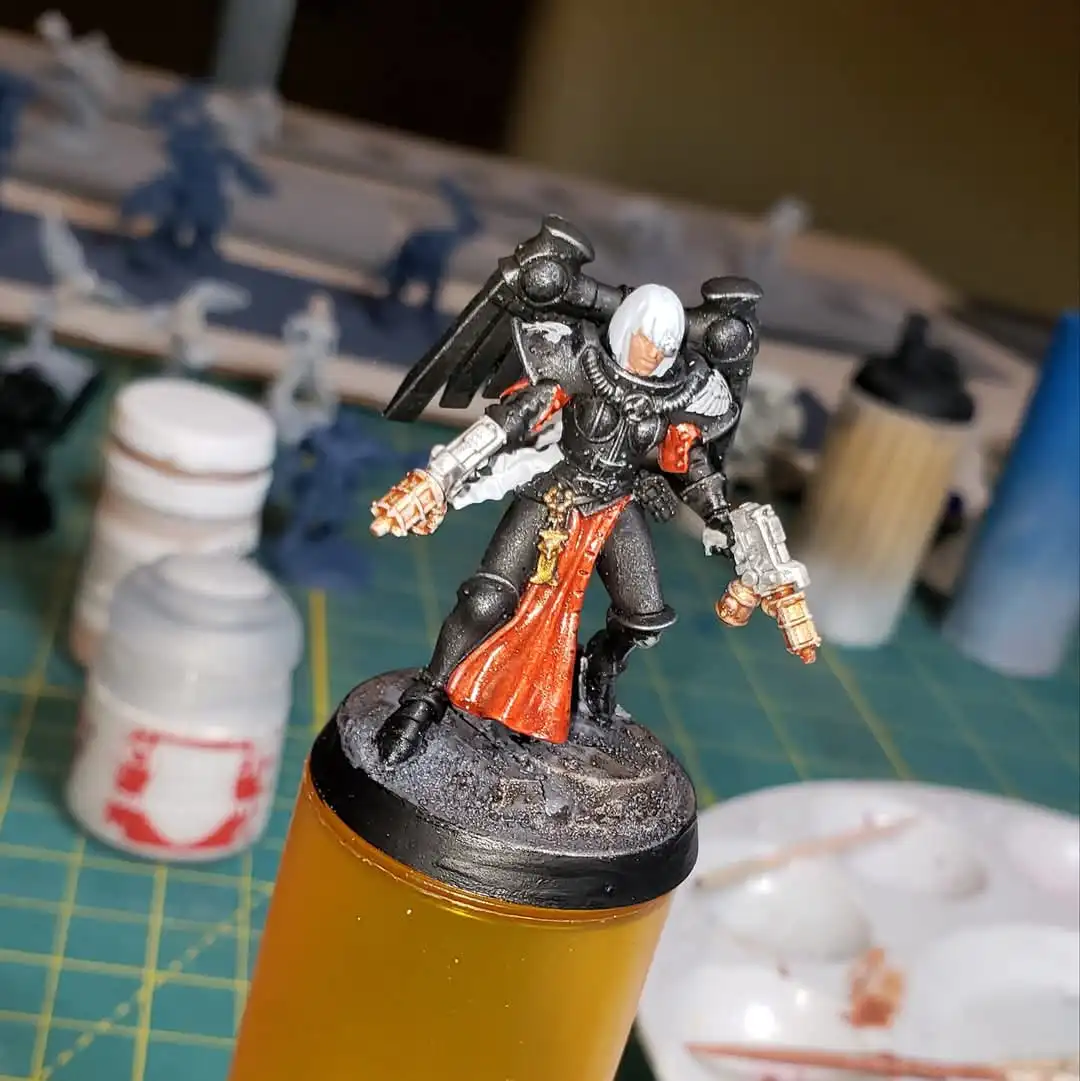 A painted miniature of a space marine with white hair, black armor, and a red cape stands on a black base atop a yellow…