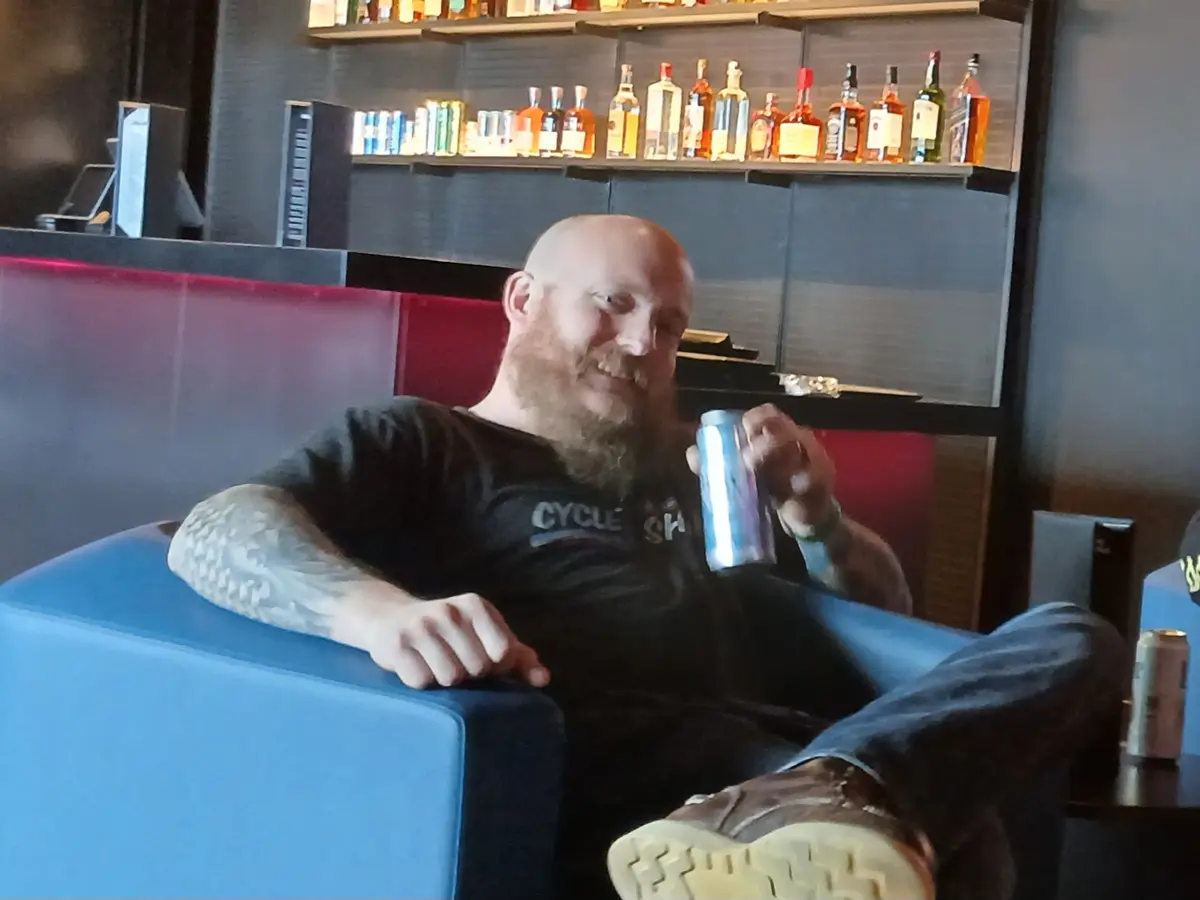 A bald man with a full beard and tattoos sits relaxed in a blue armchair in a bar, holding a silver can and smiling. Behind…