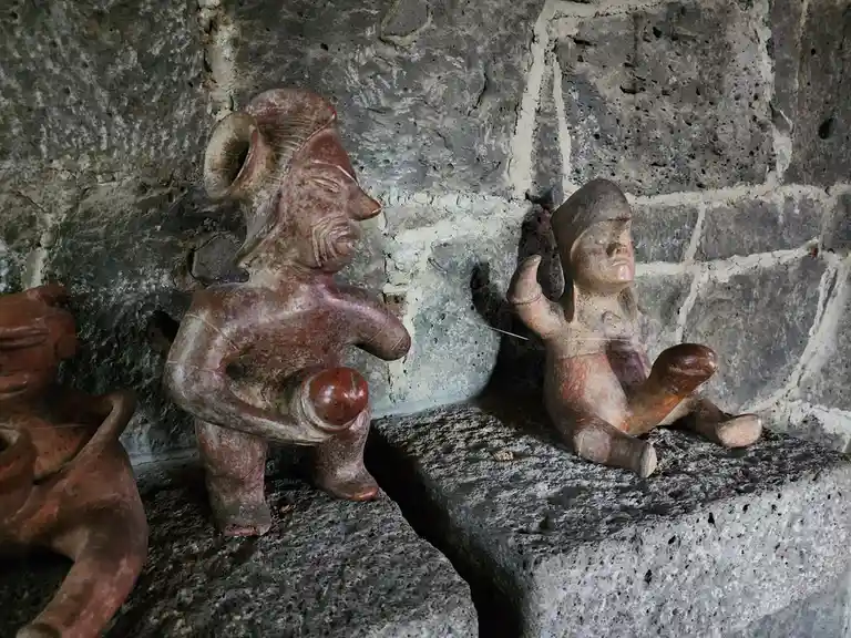 Two ancient-looking ceramic figurines sit on stone ledges against a rough stone wall; one holds a round object, the other…