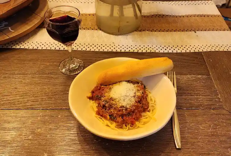 A white bowl of spaghetti topped with meat sauce and grated parmesan cheese, with a long breadstick resting on the rim…