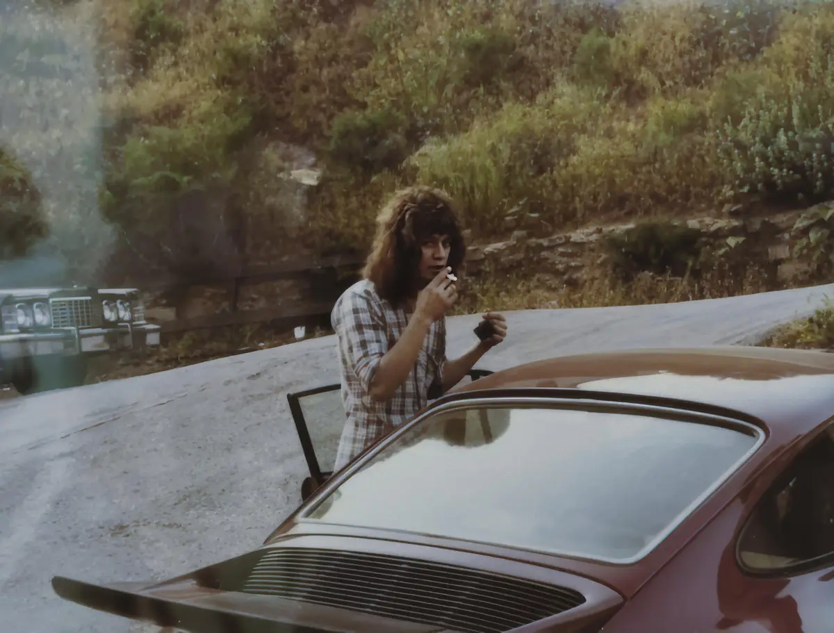 A person with shoulder-length brown hair, wearing a plaid shirt, stands beside a red car with its door open, holding a…