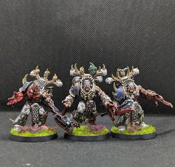 Three heavily armored, grotesque fantasy miniatures with skulls and horns stand on grassy bases against a dark textured…