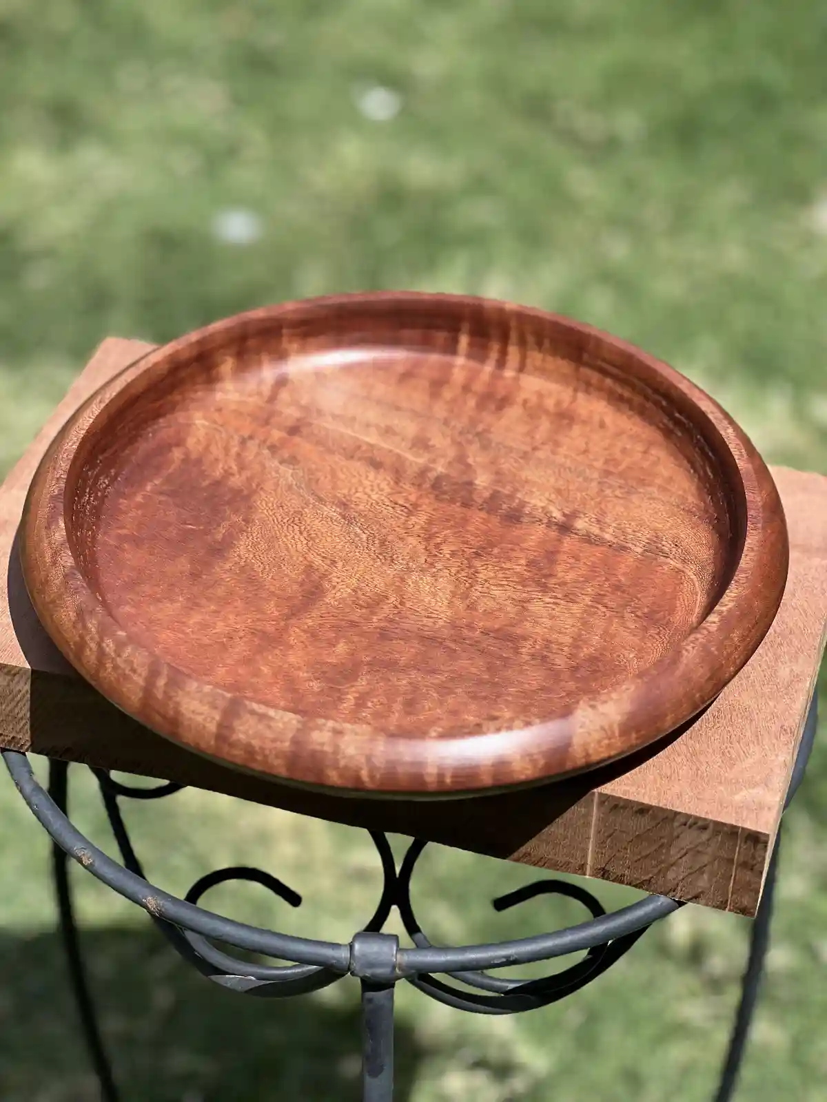 A close-up photo shows a smooth, round wooden bowl with a rich, reddish-brown finish resting on a wooden block atop a black…