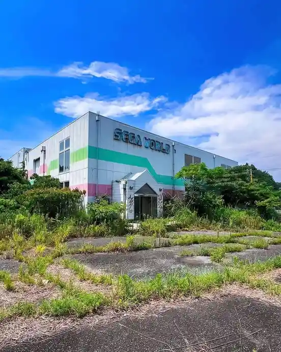 An abandoned Sega World building with overgrown weeds and grass covering the cracked pavement in front, under a bright blue…