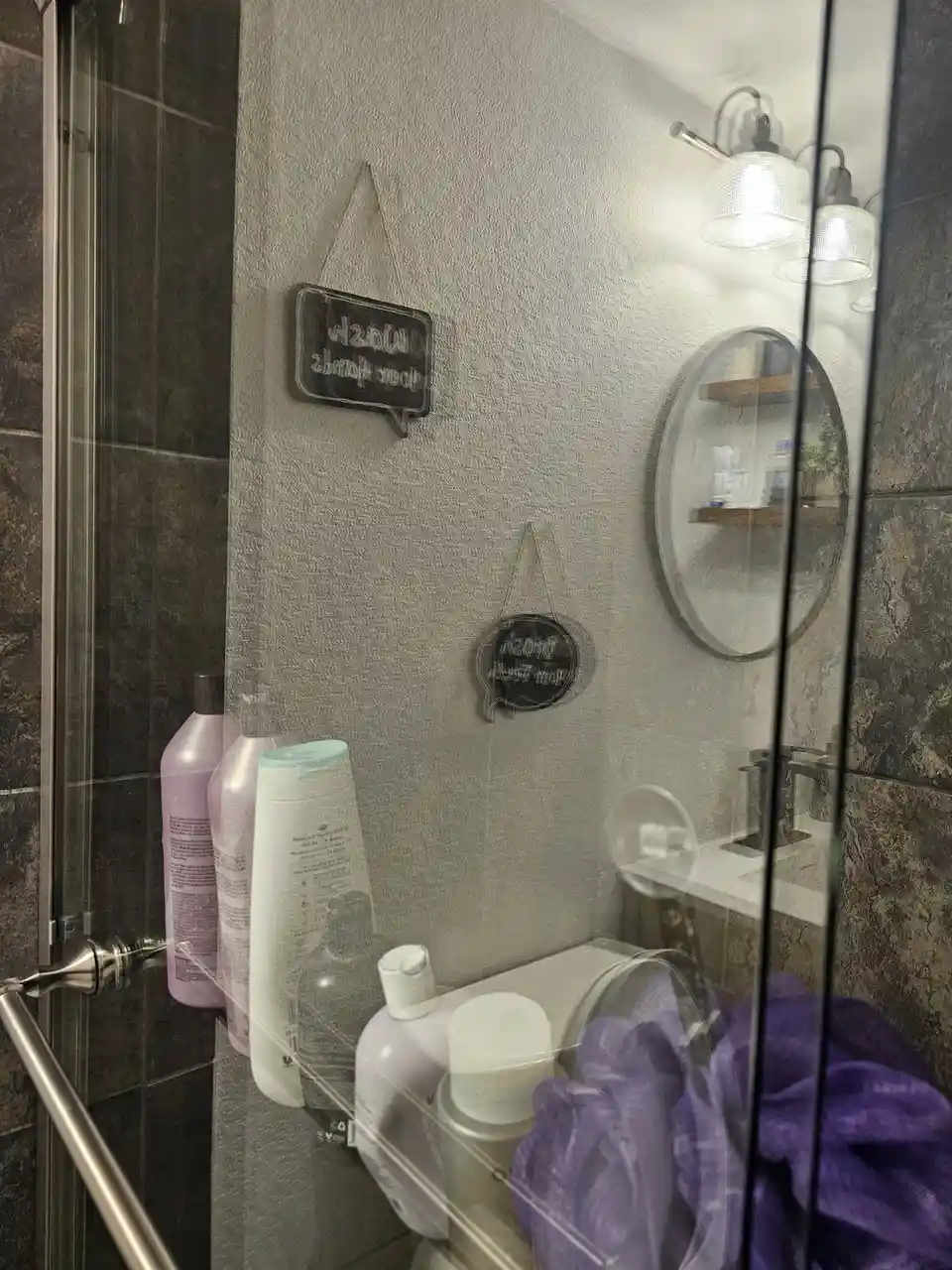 A bathroom shower enclosure with dark tiled walls and a glass door, showing various toiletries like bottles and a purple…