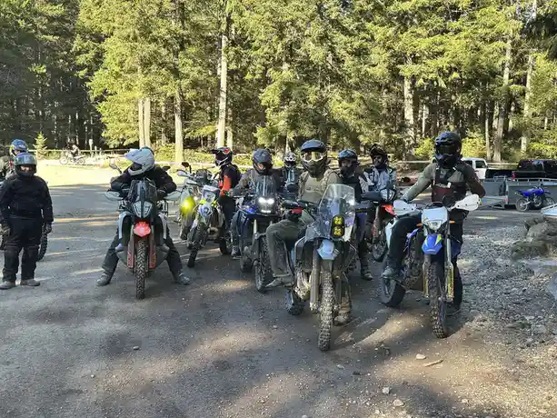 A group of motorcyclists in full protective gear, including helmets and riding suits, are lined up on dirt bikes on a gravel…