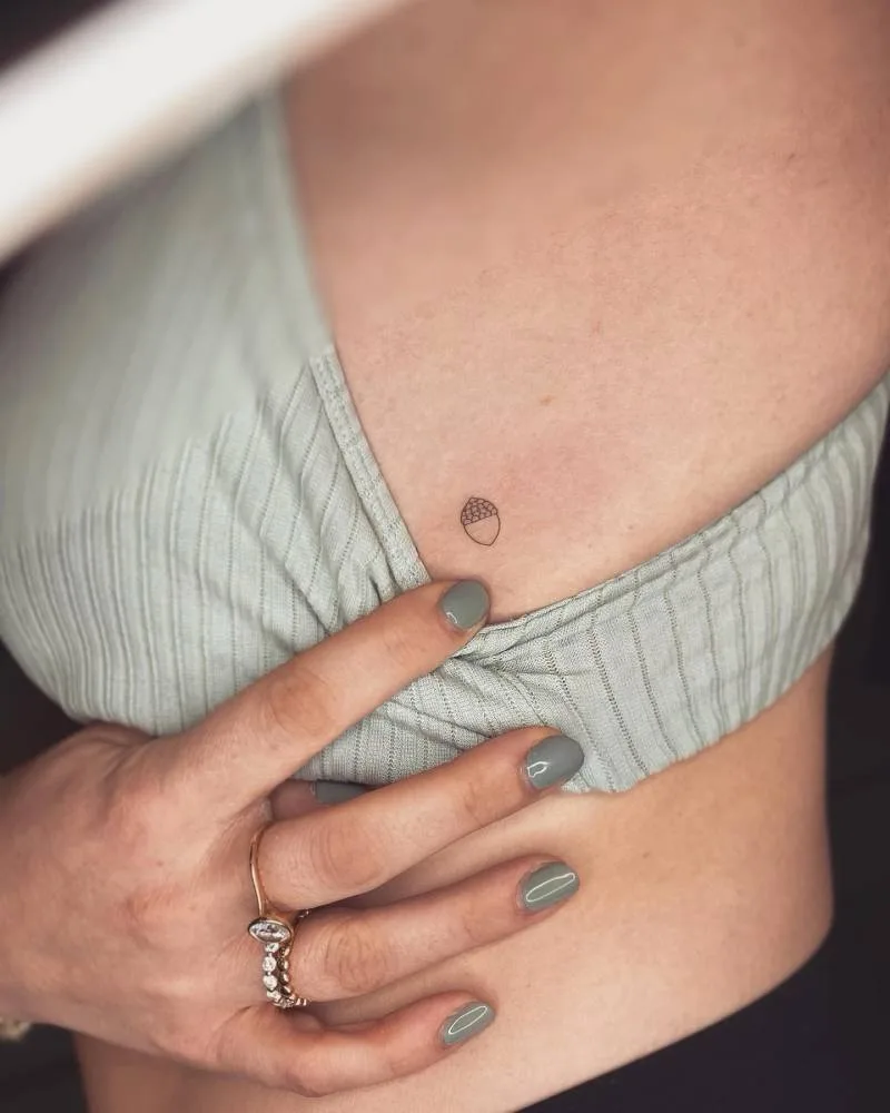 A close-up photo shows a woman's torso with a small, minimalist acorn tattoo on her ribcage, just above her bra strap. Her…