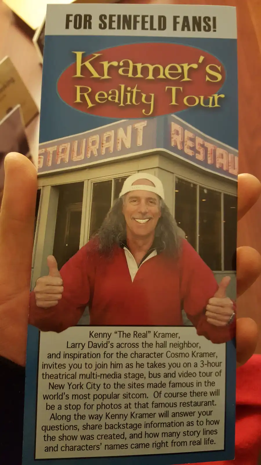 A hand holds a promotional brochure titled 'Kramer's Reality Tour' for Seinfeld fans. The brochure features a photo of Kenny…