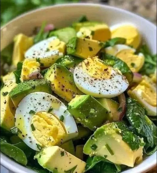 a close-up photo of a white bowl filled with a fresh green salad containing chopped avocado, sliced hard-boiled eggs…