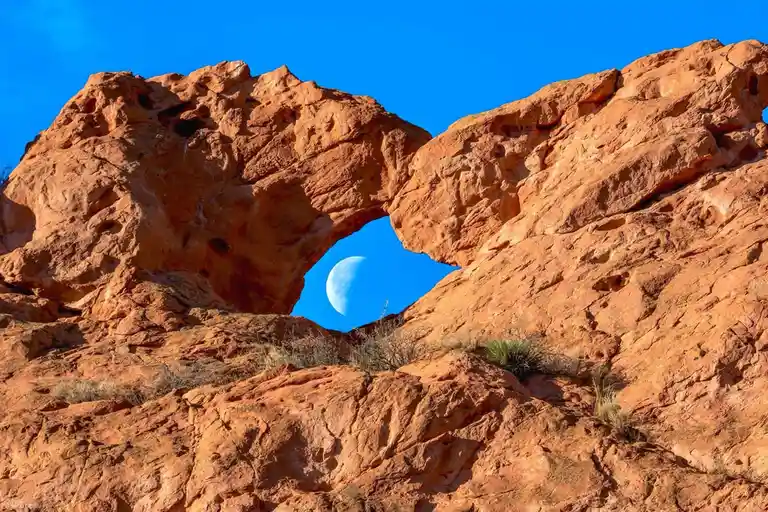 A natural rock arch in red-orange desert rock frames a crescent moon against a vivid blue sky. The overlaid text reads 'the…