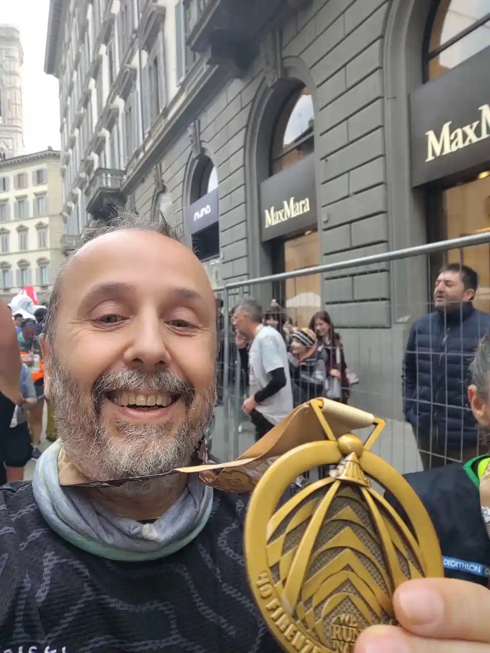 A smiling man with a gray beard and a patterned shirt holds up a large gold-colored marathon medal in front of a city street…