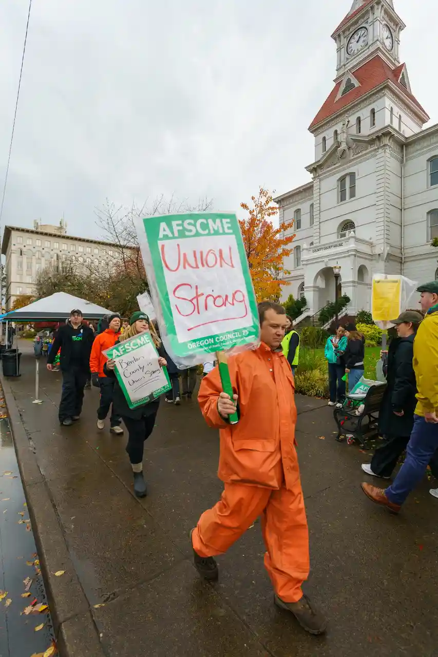 A man in a full orange jumpsuit walks in a protest march holding a green sign that reads 'AFSCME UNION STRONG OREGON COUNCIL…