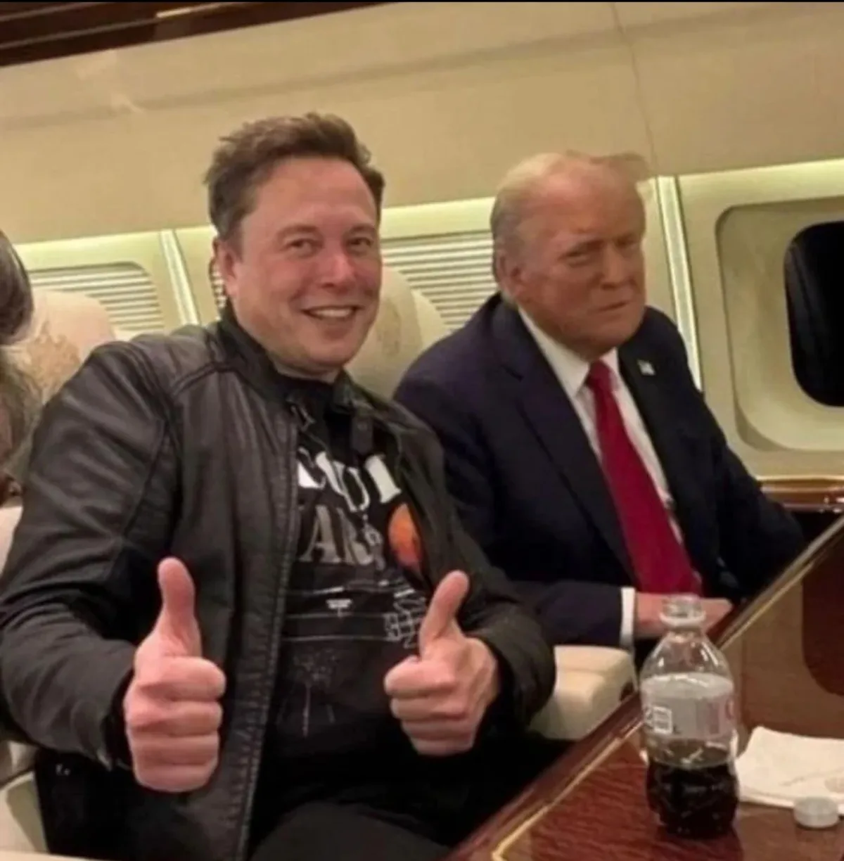 elon musk gives two enthusiastic thumbs up while sitting next to donald trump on an airplane, both men are seated at a table…