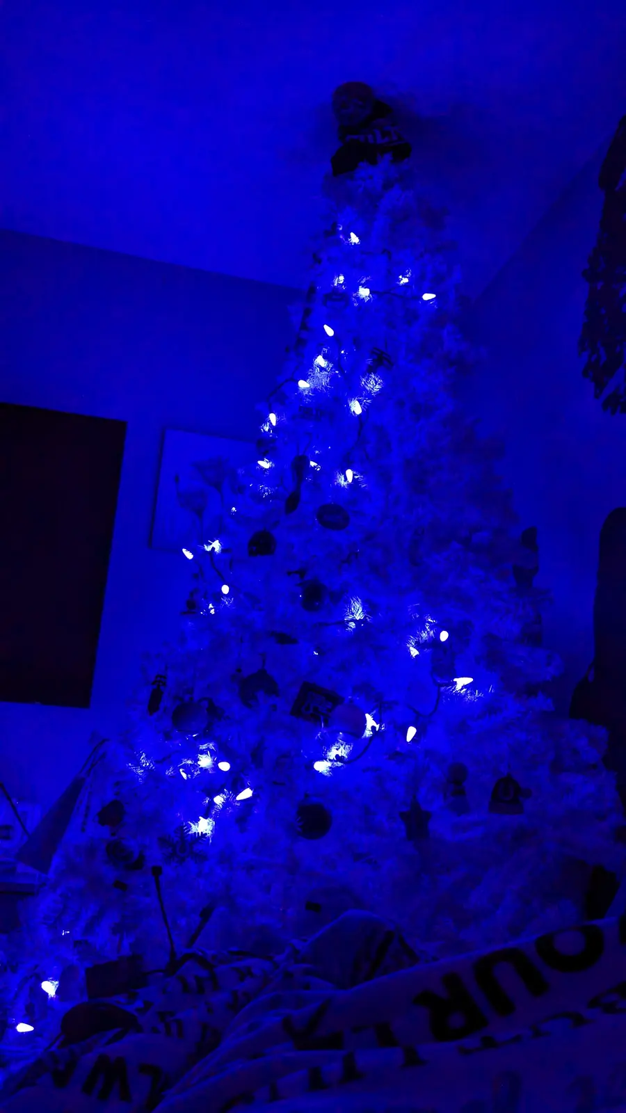 A white artificial Christmas tree is decorated with ornaments and string lights, but the entire scene is bathed in a strong…