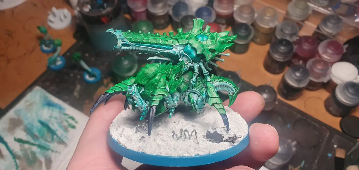 A hand holds a highly detailed, green and teal painted fantasy miniature resembling a crab-like creature with spiky armor…