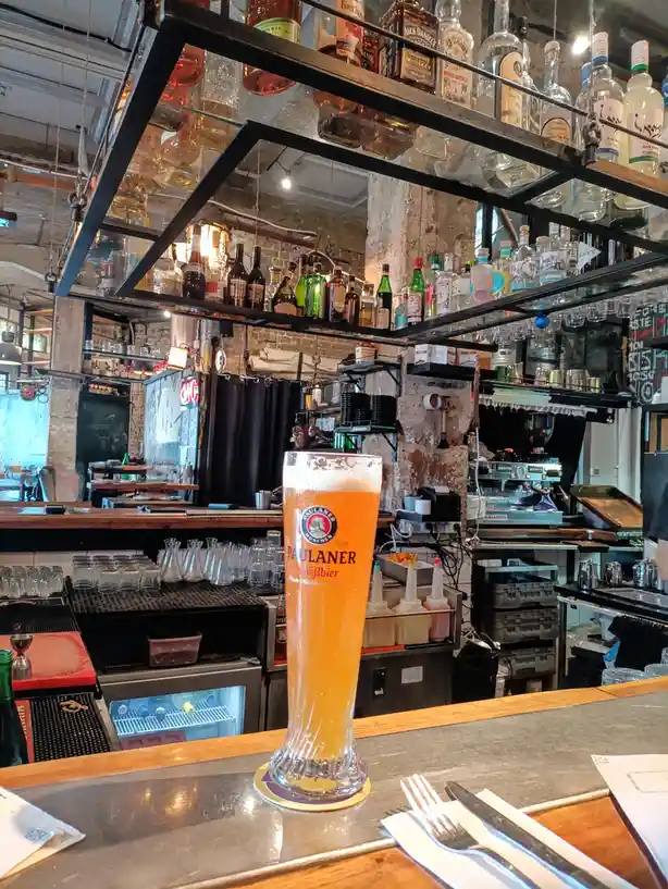 A tall glass of Paulaner Weissbier sits on a bar counter, with a backdrop of shelves stocked with various liquor bottles and…
