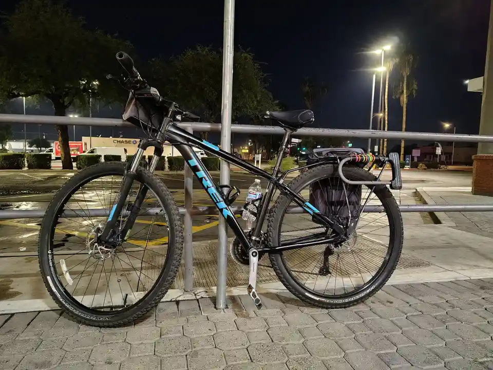A black Trek mountain bike with blue accents is parked at night against a metal railing in a wet parking lot, equipped with…