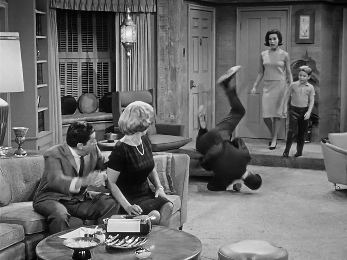 A black-and-white still from a 1960s sitcom shows a man doing a backflip in a living room while a woman and child watch from…