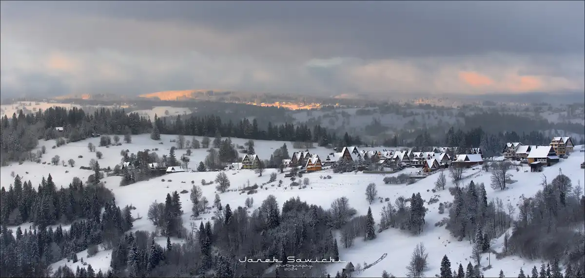 A panoramic winter landscape photograph shows a snow-covered village nestled among rolling hills and dense forests under a…