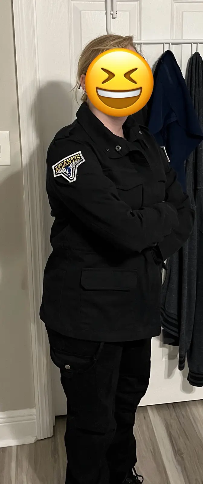 A person stands with arms crossed wearing a black uniform with an 'Atlantis' patch on the left sleeve, their face obscured…