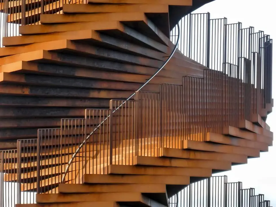 A close-up photograph of a large, rust-colored spiral staircase with metal railings, viewed from below against a bright sky…