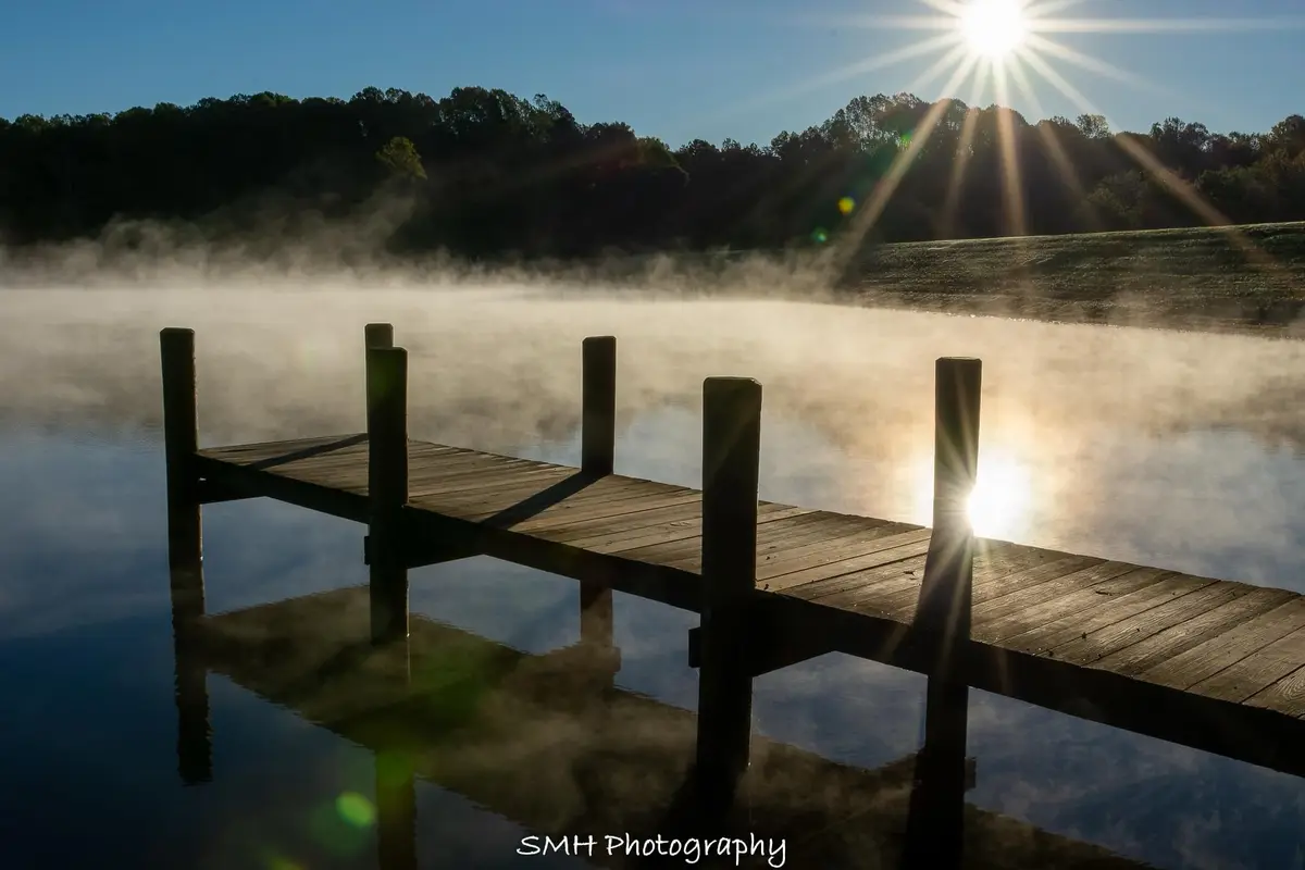 A serene photograph of a wooden dock extending over a misty lake at sunrise, with the sun casting bright rays through the…