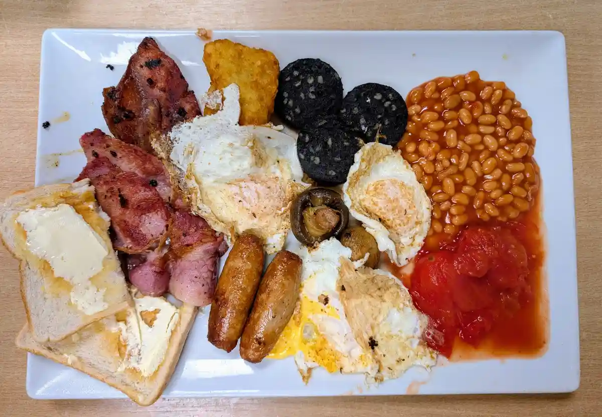 A white plate holds a full English breakfast with fried eggs, bacon, sausages, black pudding, baked beans, grilled tomatoes…